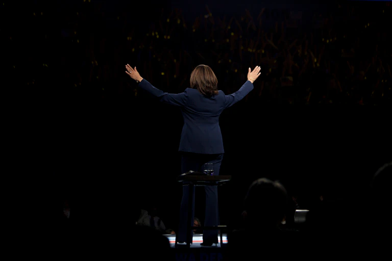 Kamala Harris de espaldas y los brazos abiertos sobre fondo negro.