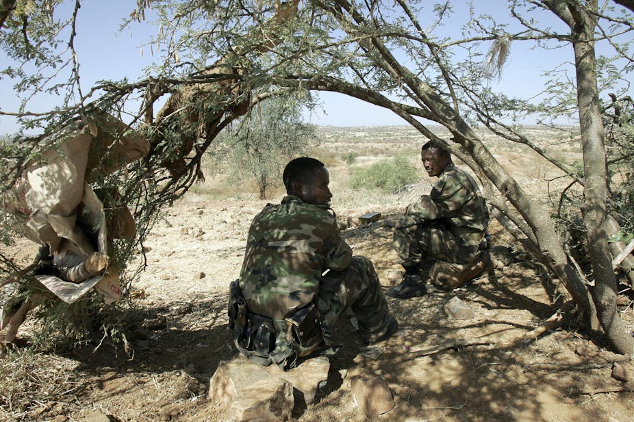 Two soldiers sit in the patchy shade of a tree. They are dressed in military camouflage uniforms with a vast plain of veld in the distance.