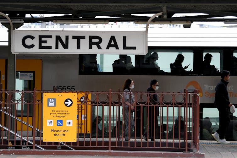 Two people wearing face masks walk down the platform of a station in front of a crowded train.