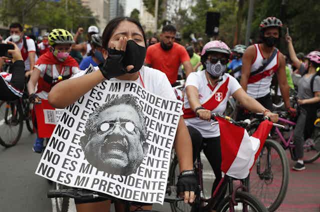 Peruvian woman riding a bicycle and carrying anti-government sign flanked by other protesters on cycles wearing Peru's national colours.