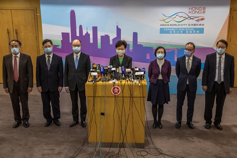 Carrie Lam stands at podium with microphones surrounded by six colleagues, all wearing masks.