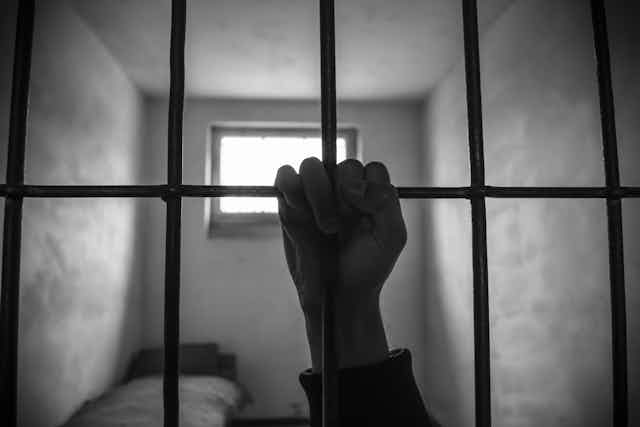 A black and white photograph of a prison cell with the silhouette of a hand grasping bars