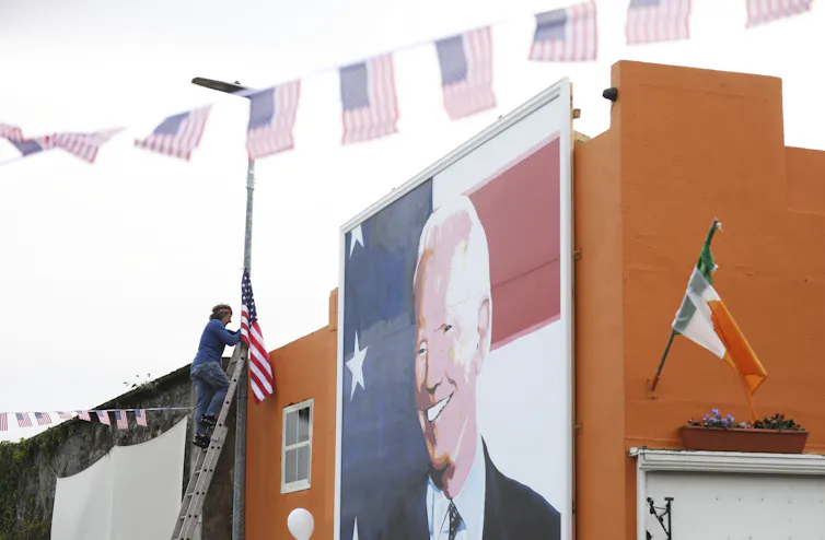 A man stands on a ladder next to a mural of Joe Biden.