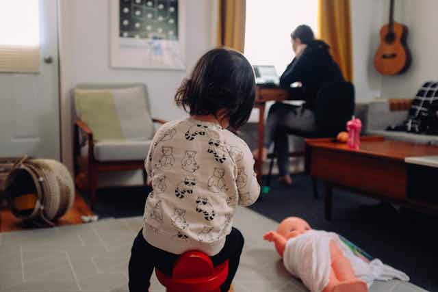 A mother works on her laptop while her child is playing in the foreground of their living room