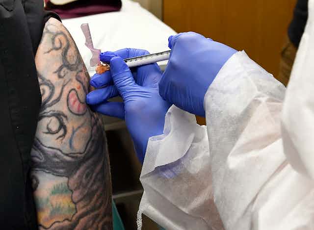 The gloved hands of a health-care worker giving an injection on a tattooed upper arm