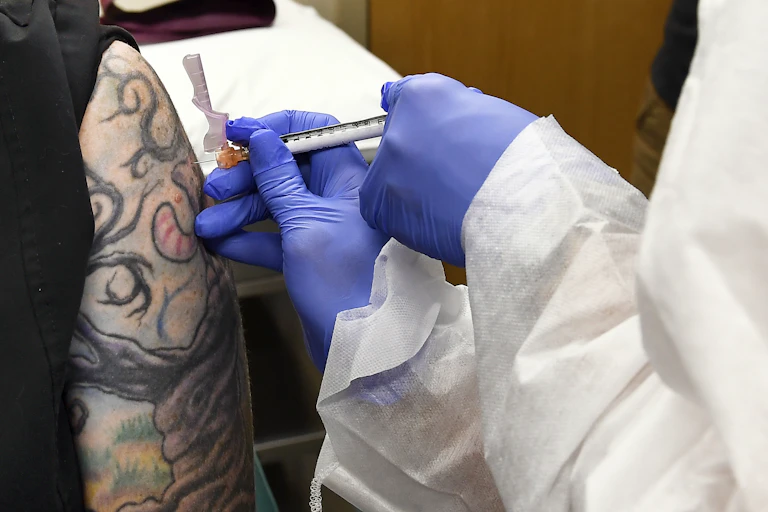 The gloved hands of a health-care worker giving an injection on a tattooed upper arm