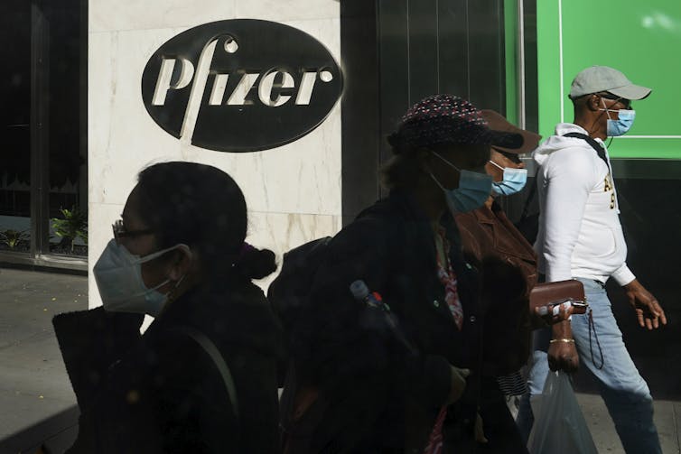 People wearing face masks walk past a Pfizer sign at street level.