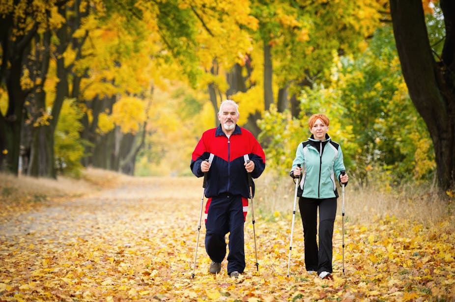 Middle-aged couple using nordic walking sticks to walk down a leaf-covered autumn path.