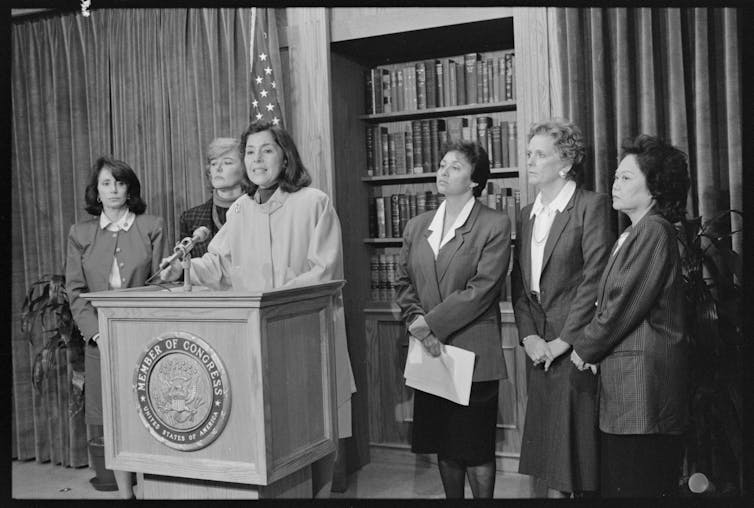 Black and white image of six congresswomen in suits standing at a lectern