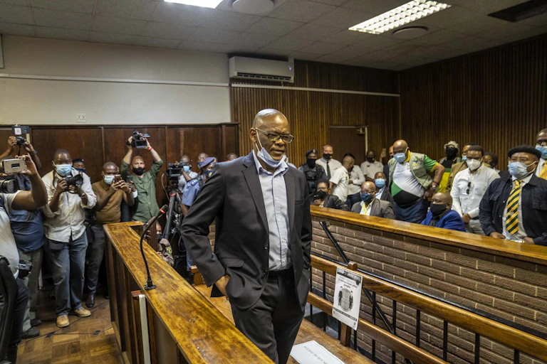 A thin bespectacled man wearing a dark suit, blue shirt with an open collar and a Covid-19 mask below his mouth leaves court room