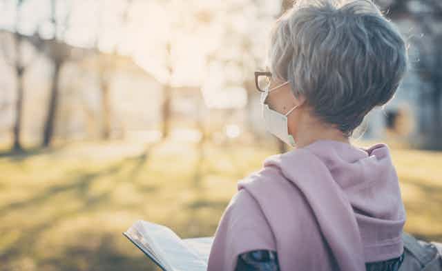 Una mujer mayor, con el pelo corto y gris y con gafas y mascarilla, lee sola en un jardín.