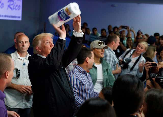 Donald Trump hold a paper towel roll like a football ready to toss to the crowd. Journalists look on. 