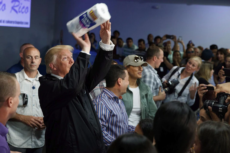 Donald Trump hold a paper towel roll like a football ready to toss to the crowd. Journalists look on.