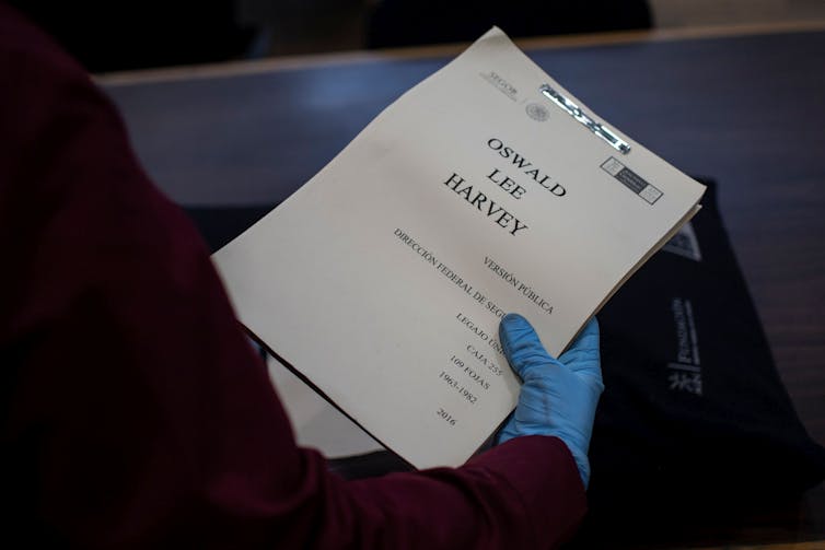 Gloved hand holds a paper report entitled 'Lee Harvey Oswald'