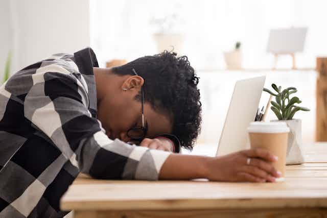 A young African American woman lies her head down on a her desk while holding a cup of coffee next to her laptop.
