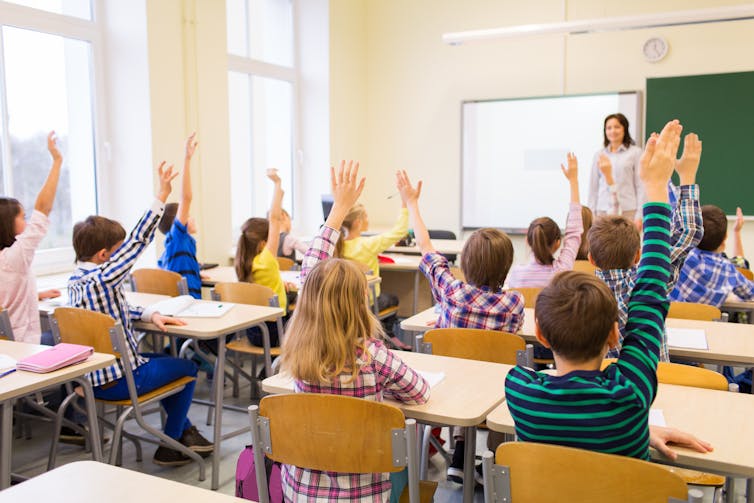 Students sitting at rows of desks facing the teacher, with their hands up.