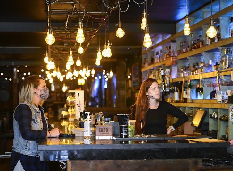 A woman wearing a mask sits at a bar while a bartender stands behind it. Both look at a TV set.