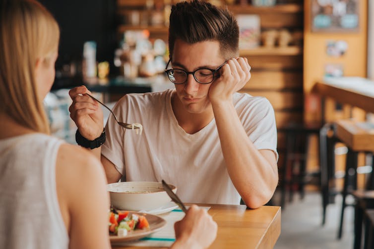 A man and a woman are eating in a cafe, but the man is disinterested in his food.