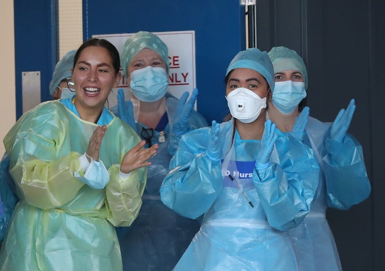 Four women in personal protective equipment clap at a hospital door.