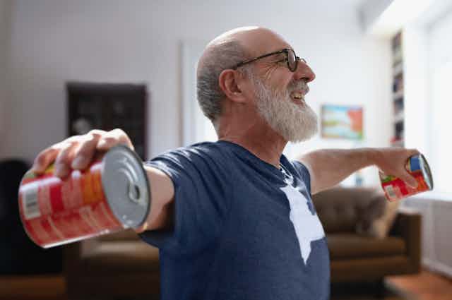 Older man excercising from home by lifting two cans of tomatoes up to shoulder height.