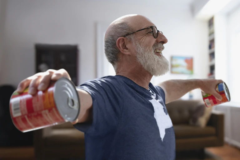 Older man excercising from home by lifting two cans of tomatoes up to shoulder height.