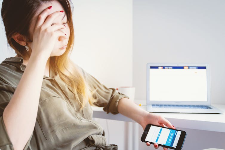 A young woman sitting in front a desk and laptop, looking at her phone with her face in her hand.