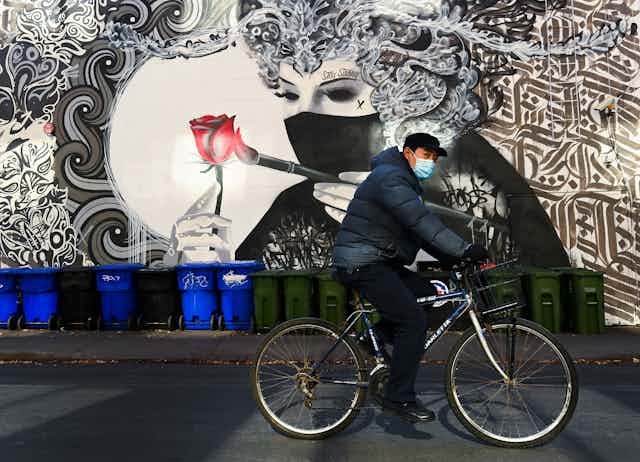 A man wearing a face mask rides a bike past a mural of a woman wearing a face mask