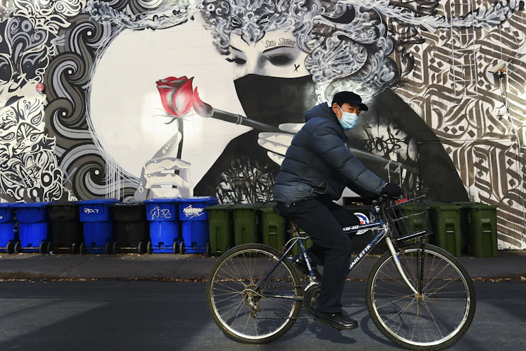 A man wearing a face mask rides a bike past a mural of a woman wearing a face mask