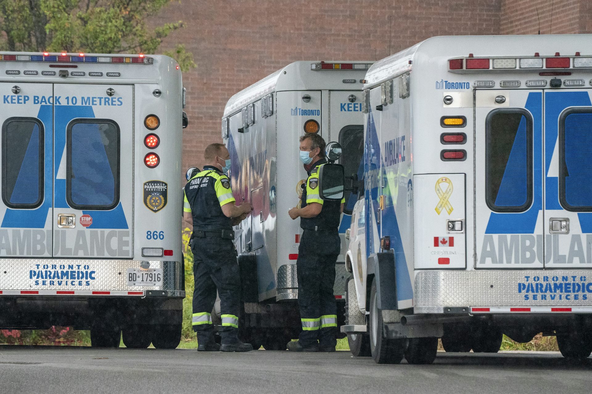 Two paramedics wearing masks stand talking between ambulances.
