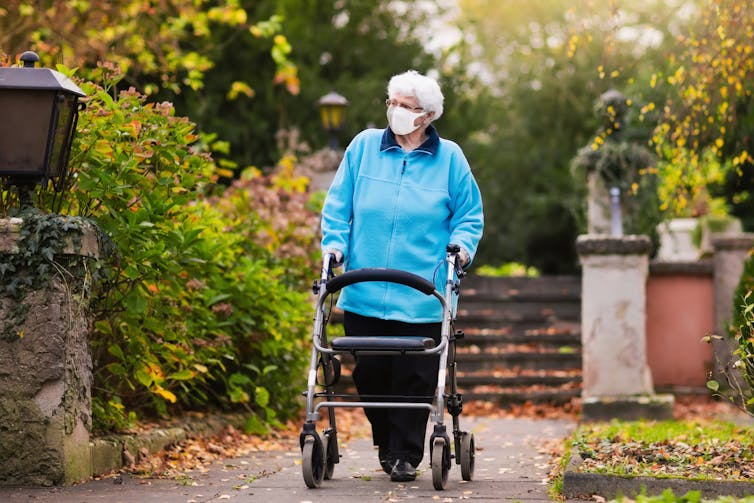 An elderly lady wearing a mask walks with a frame in a garden.