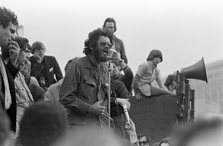 Archival photo of man in front of microphone with small crowd behind him