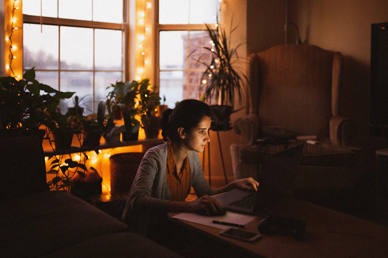 A woman sits in front of her laptop in a dimly lit apartment with a wing chair and houseplants behind her.
