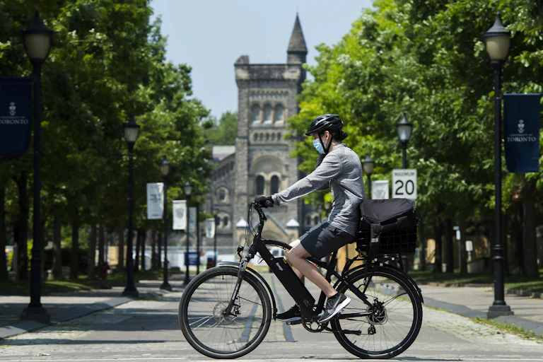 A person in a face mask and bike helmet rides a bicycle past a university building