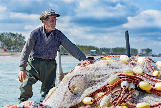 An older fisherman stands next to a pile of nets