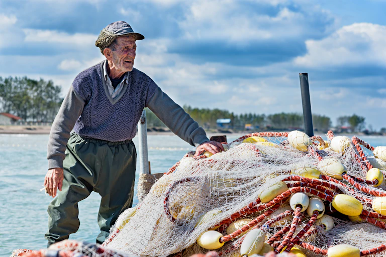 An older fisherman stands next to a pile of nets