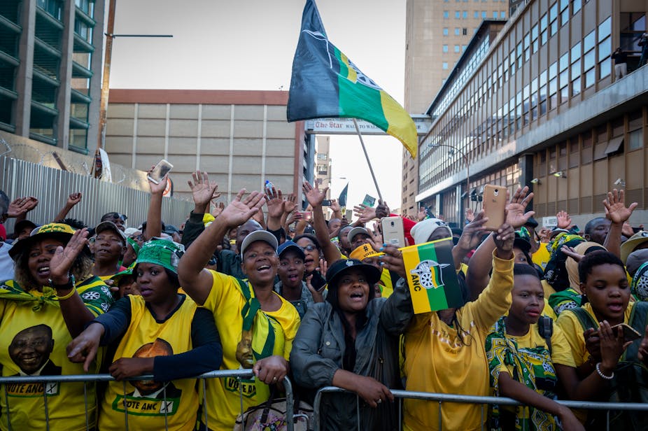 Crowd of people in the street, wearing green and yellow shirts and holding party flags, arms raised