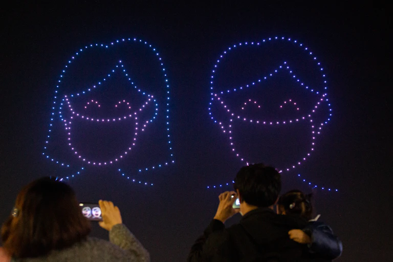 Drones form a picture in the night sky of a woman and a man wearing masks.