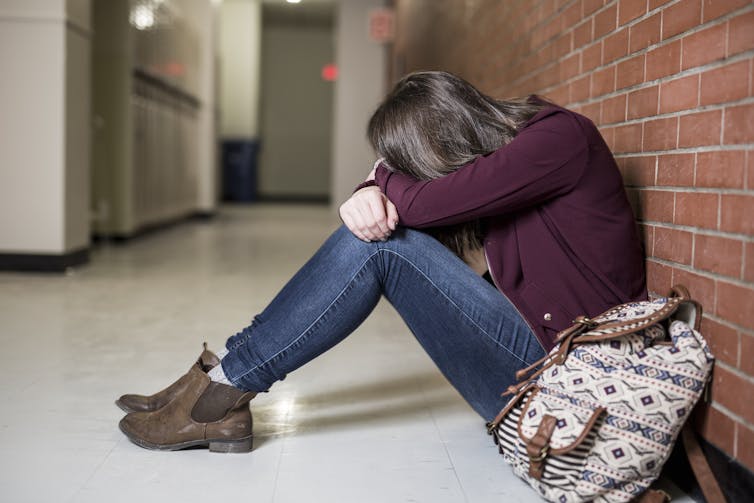A teenage on the ground near lockers with her head on her knees.