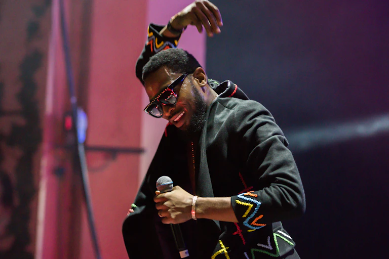 A man in shades holds a hand above his head and a microphone in his hand, clearly performing on stage.