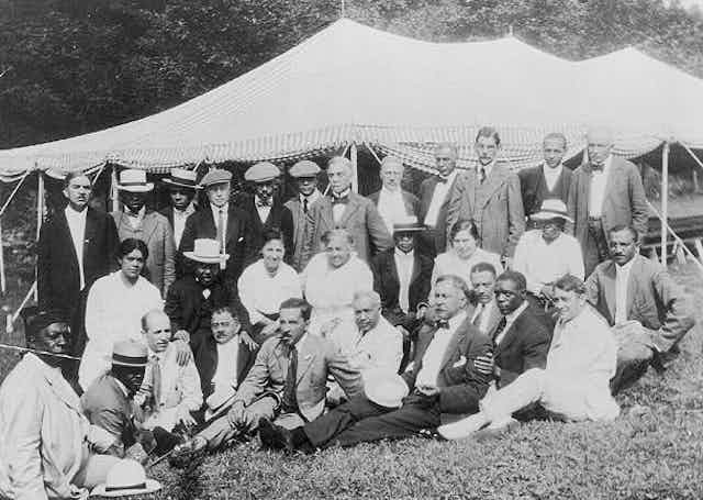 A group of people gather outdoors, in front of a tent, in an old photo.