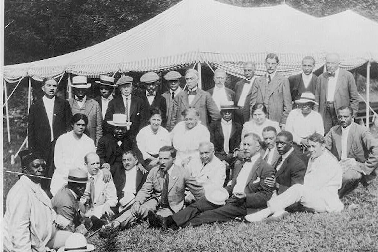 A group of people gather outdoors, in front of a tent, in an old photo.