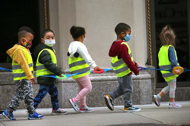 Five small children wearing face masks and yellow vests walk in single file holding a strap