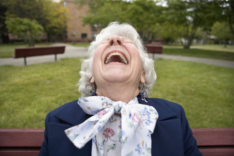 Femme aux cheveux blancs riant sur un banc public dans un parc