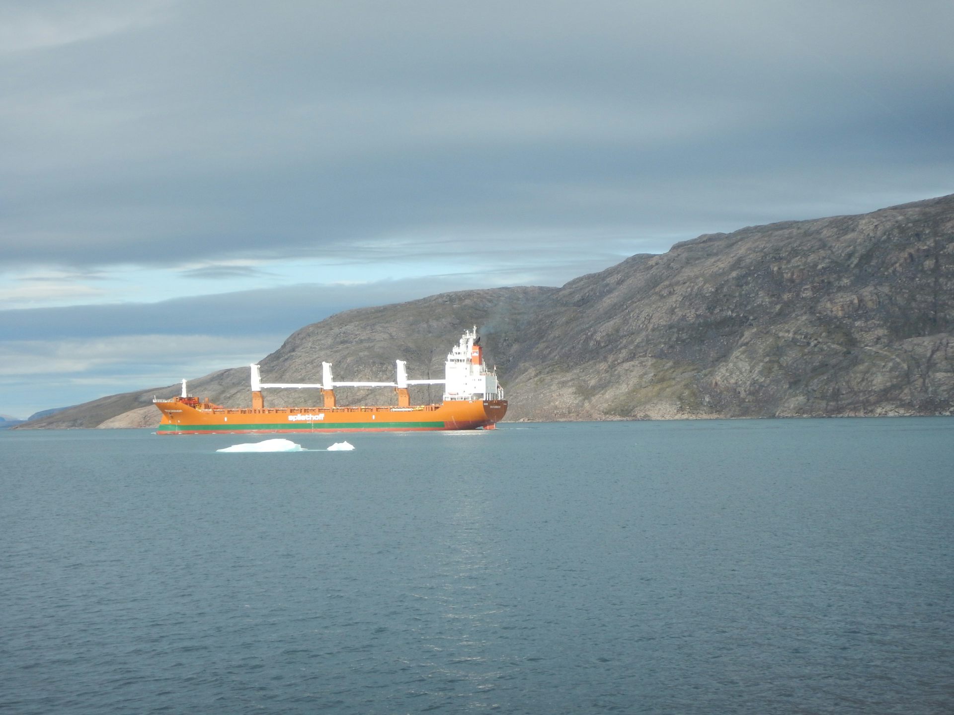 An orange ship sits in icy water with a rocky slope behind it.