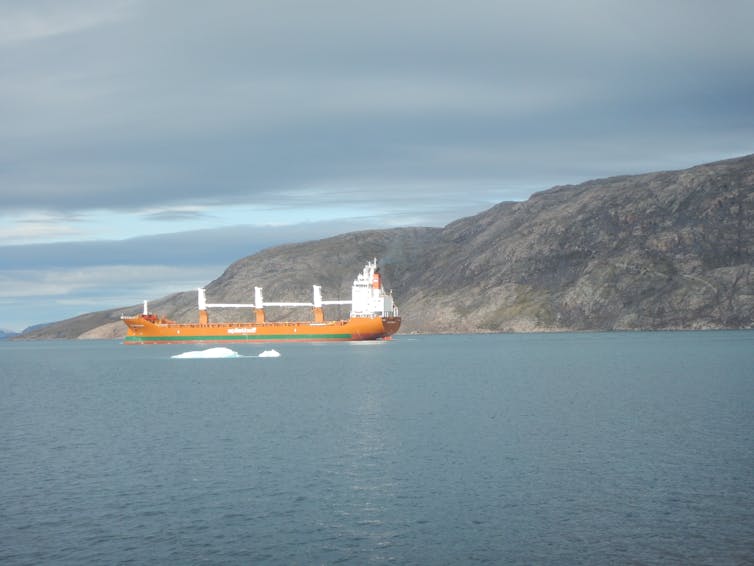An orange ship sits in icy water with a rocky slope behind it.