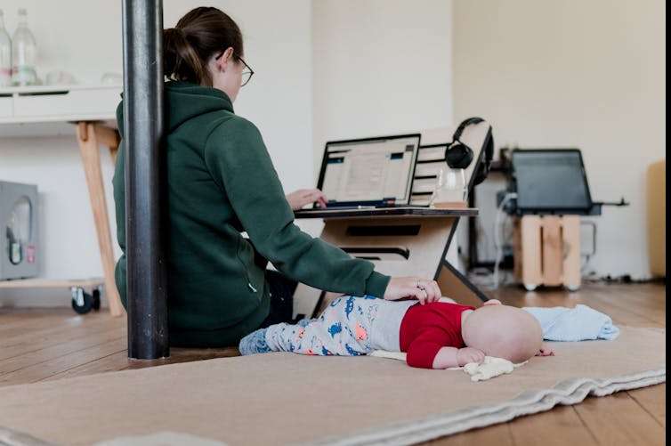Mother seated on floor and comforting baby while working at laptop