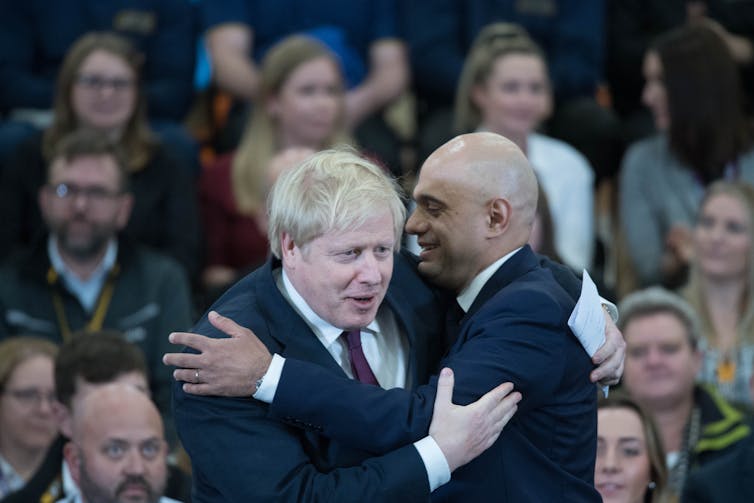 Boris Johnson and Sajid Javid embracing in front of seated audience.