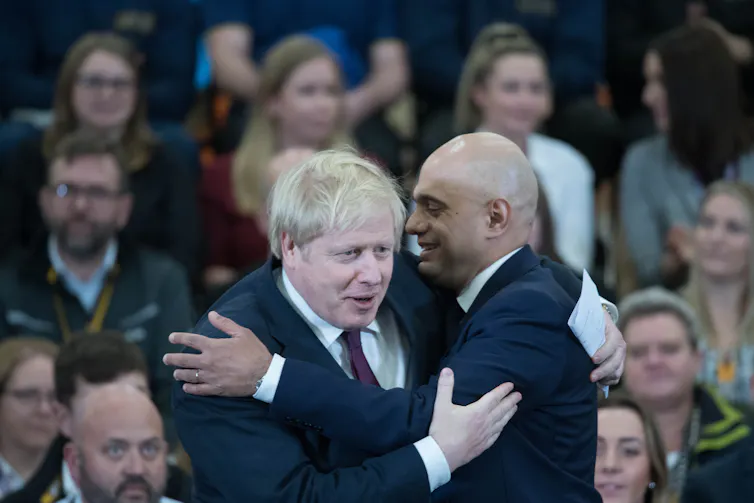 Boris Johnson and Sajid Javid embracing in front of seated audience.