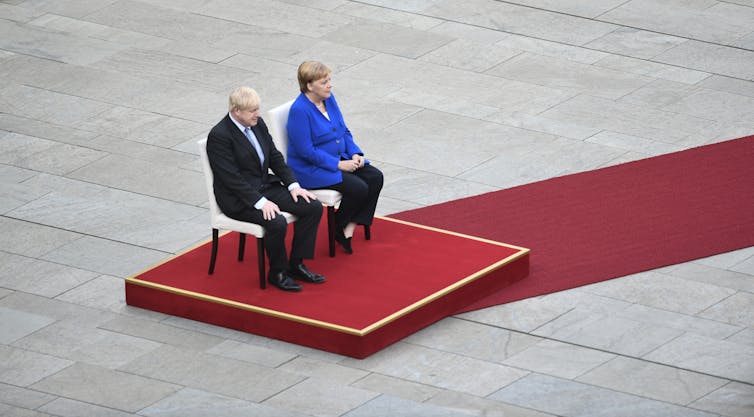 Boris Johnson and Angela Merkel sitting on a podium.