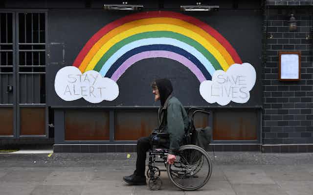 A man in a wheelchair passes a street mural of a rainbow and the slogan "stay alert save lives".